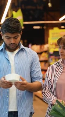 Genç Hintli çift markette yiyecek alışverişi yapıyor. Elinde bir ürün tutan ve alışveriş yapmadan önce ortağına danışıyor. O da alışveriş arabasında taze pırasa tutuyor.