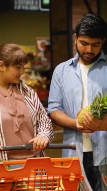 Genç çift gülümsüyor ve konuşuyor. Bir marketin sebze reyonundan taze bir ananas seçerken sağlıklı meyveyi alışverişe devam etmeden önce turuncu alışveriş arabalarına koyuyorlar.