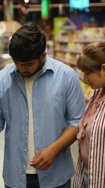 Düşünceli Hintli erkek ve kadın, büyük, modern bir süpermarketin koridorlarında birlikte alışveriş yaparken, bilinçli bir seçim yaparak, bir ev kimyasal ürününün etiketini dikkatle okuyorlar.