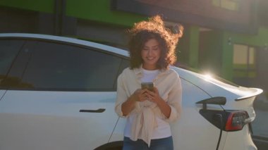 Neşeli, kıvırcık saçlı genç bir kadın modern elektrikli aracına yaslanıyor ve elektrik santralinde şarj olurken akıllı telefon kullanıyor, güneşli havanın tadını çıkarıyor ve bağlı kalıyor.