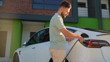 Yakışıklı sakallı bir adam modern evinin önünde beyaz elektrikli aracını şarj etmek için elektrik kablosu takıyor ve akü durumu için akıllı telefonunu kontrol ediyor.