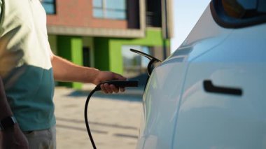 Arka planda yeşil enerji ve çevre dostu taşımacılığı destekleyen modern bir konut binası olan şarj için elektrikli bir araca elektrik kablosu yerleştiren bir adamın elini kapatın.