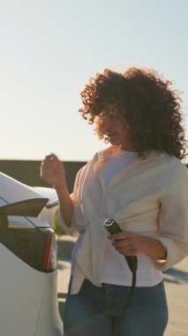 Kıvırcık saçlı bir kadın şarj istasyonunda modern elektrikli aracını fişe takıyor ve sonra pilin şarj olmasını beklerken bir telefon görüşmesi yapıyor.