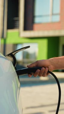 Bir adamın şarj aletini modern beyaz elektrikli bir araca takıp sürdürülebilir ve çevre dostu bir taşımacılık için temiz ve yenilenebilir enerjiyle yakıt ikmali yaptığı görüntüler.