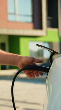 Beyaz bir elektrikli arabanın şarj prizini söken bir adamın eli kapalı. Bu da güneşli bir günde bir konut istasyonunda tam akü şarjının bittiğini gösterir.