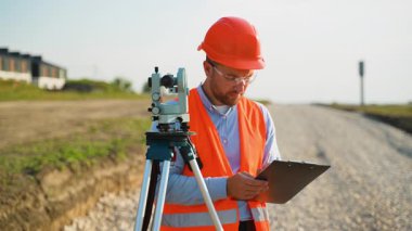 Uzman bir araştırmacı, tripodun üzerinde bir teodolitle çalışıyor. Ölçümleri alıyor ve bir yol inşaatında bir panoya yazı yazıyor. Üzerinde sıkı bir şapka ve yüksek görüş mesafesi yeleği var.