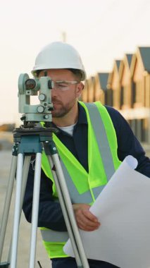 Açık hava endüstriyel şantiyesinde çalışırken, bir teodolit kullanarak ve bir planı analiz ederek, sert şapkalı ve yüksek görüş açısına sahip beyaz erkek araştırmacı.