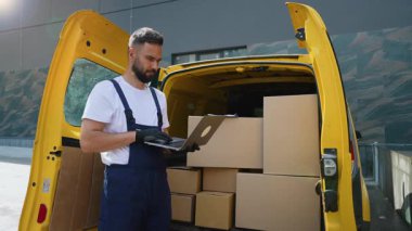Ciddi bir teslimat işçisi, karton kutularla dolu sarı bir minibüsün yanında duruyor ve envanter listesini kontrol etmek için dizüstü bilgisayar kullanıyor, ticari bir ulaşım servisi için sevkiyat ve lojistiği yönetiyor.