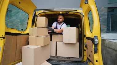 Sarı bir minibüsün arkasında oturan, karton kutularla çevrili genç bir kurye gülümsemeden ve kameraya işaret vermeden önce panosundaki listeyi kontrol ediyor.