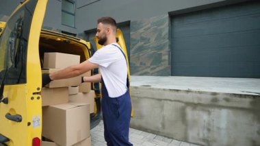 Mavi üniformalı arkadaş canlısı kurye sarı ticari bir araçtan karton bir kutuyu indiriyor, kameraya dönüyor ve gülümsüyor, çevrimiçi alışveriş için hızlı ve güvenilir nakliye hizmetleri sunuyor.