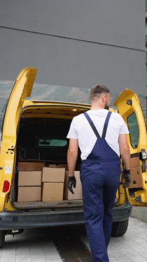 Mavi tulumlu ve eldivenli profesyonel kurye, karton kutu ve paketleri nakliye ve taşımacılık hizmetlerine hazırlanmak üzere sarı bir minibüsün arkasına dikkatlice yüklüyor.