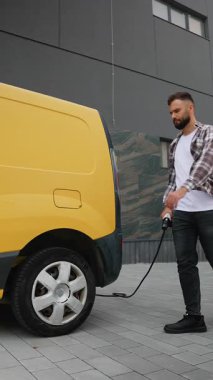 Genç adam sarı elektrik dağıtım minibüsüne şarj kablosu bağlayıp şehrin modern şarj istasyonunda aracın şarj olmasını beklerken akıllı telefonunu kullanıyor.