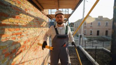 Bitmemiş tuğla bir binanın yanında iskele üzerinde durmuş kameraya bakıp gülümserken ruh seviyesinde bir aletle dans eden sağlam şapkalı ve tulumlu olumlu bir genç.