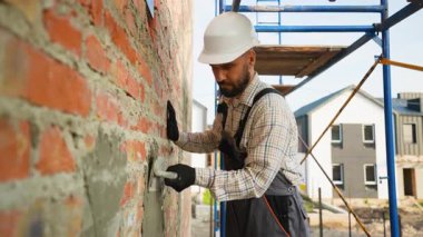 Sert bir şapkayla konsantre olmuş erkek müteahhit titizlikle dış tuğla duvara bir malayla yeni alçı yapıştırıyor ve bir inşaat alanında iskele üzerinde çalışıyor.