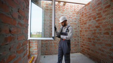 Sabit şapkalı ve üniformalı profesyonel müteahhit titizlikle tamamlanmamış kırmızı tuğladan duvarları olan yeni bir binaya modern plastik bir pencere yerleştiriyor.