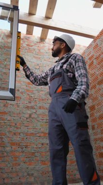 İnşaat alanındaki tuğla bir duvara yeni bir pencere çerçevesi monte etmek için ruh seviyesinde bir araç kullanan sert şapka ve tulum giyen profesyonel bir inşaatçı.