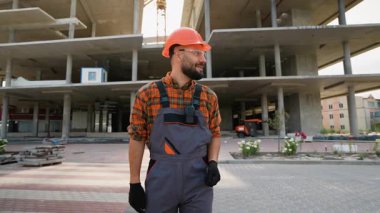 Sert şapkalı, üniformalı, neşeli bir şekilde dans eden, bitmemiş beton bir binanın önünde kameraya gülümseyen, başarılı projeyi kutlayan pozitif sakallı inşaat işçisi.