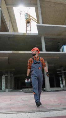 Kendine güvenen erkek müteahhit, sabit bir şapka takıyor ve inşaat halindeki bir binada yürüyor, arkasında parlak gökyüzüne karşı görünen bir vinç ve çekiç taşıyor.