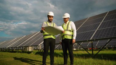 İki erkek mühendis güneş enerjisi santralinde dikilip bulutlu bir gökyüzü ve yeşil çimenlere karşı yenilenebilir enerji projesinin planını tartışıyor.