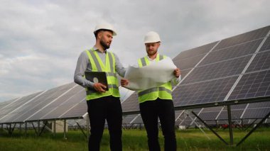 Güneş çiftliğinde dikilirken bir proje planını tartışan miğfer ve güvenlik yelekleri giyen iki mühendis, biri dizüstü bilgisayar ve diğeri de fotovoltaik panelleri teftiş eden bir plan tutuyor.