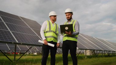 İki erkek elektrik mühendisi, yenilenebilir enerji santralinde güneş panelleri arasında dururken dizüstü bilgisayar ve planlar kullanarak bir projede işbirliği yapıyorlar.