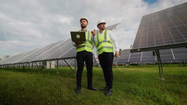 Güvenlik yelekleri ve başlıklarda iki erkek enerji uzmanı fotovoltaik paneller arasında dizüstü bilgisayarla bir projeyi tartışıyorlar, takım çalışması ve sürdürülebilir güç yeniliği sergiliyorlar.