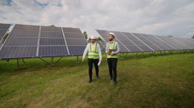Güneş çiftliğinde yürüyen iki erkek mühendis, fotovoltaik panelleri temiz, sürdürülebilir ve alternatif enerji üretimi için incelerken projeyi tartışıyorlar.