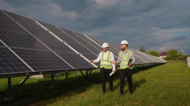 Güvenlik yelekleri ve başlıklı iki profesyonel erkek mühendis bir elektrik santralinde bir sıra güneş paneli boyunca yürürken, bir tablet ve planlar tutarken kurulum projesini tartışıyorlar.