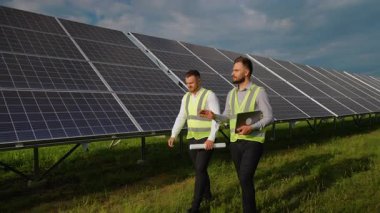 Güvenlik yelekleri giymiş iki erkek ekoloji mühendisi fotovoltaik paneller arasında yürürken, biri dizüstü bilgisayar, diğeri de planları elinde tutarken yenilenebilir enerji projesini tartışıyorlar.
