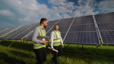 Güneş çiftliğinde yürüyen güvenlik yelekleri giyen iki erkek mühendis, projeyi bir dizüstü bilgisayar ve planlarla tartışıyor, yenilenebilir enerji üretimi için fotovoltaik panelleri kontrol ediyorlar.