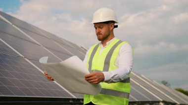 Sağlam bir şapka ve güvenlik yeleği giymiş kendinden emin erkek mühendis yenilenebilir enerji projesinin planını analiz ediyor. Bulutlu bir gökyüzünün altında büyük bir güneş paneli kurulumunun önünde duruyor.