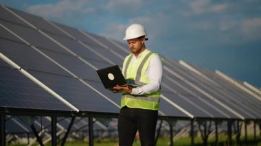 Kasklı ve üniformalı genç erkek mühendis bilgisayarını açıp fotovoltaik bir istasyonda güneş panelinin verimliliğini ölçerek yenilenebilir temiz enerji üretimini sağlıyor.