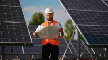Bir elektrik santralinde güneş panelleri arasında duran sert şapka ve güvenlik yeleği giyen beyaz erkek mühendis proje planlarını dikkatle gözden geçiriyor ve ekipmanları denetliyor.