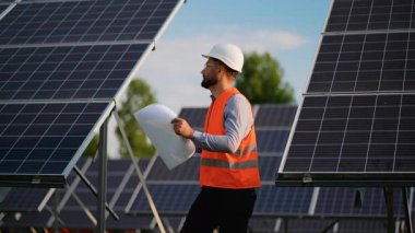 Bir güneş enerjisi santralindeki fotovoltaik panellerin montajı sırasında elinde bir proje olan sabit şapkalı erkek mühendis, yenilenebilir enerji projesinin ilerlemesini değerlendiriyor.