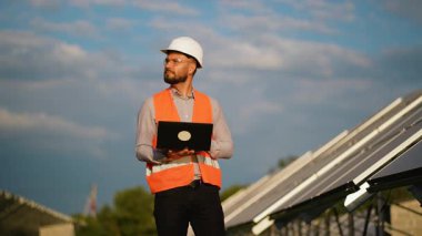 Bir güneş çiftliğinin bakım kontrolü için dizüstü bilgisayar kullanan ve arka planda güneş panelleri olan kameraya gülümseyen kasklı ve güvenlik yeleği giyen kendinden emin bir erkek mühendis.