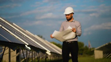 Yenilenebilir enerji çiftliğinde güneş panelleri arasında dururken planlarını inceleyen kendinden emin erkek mühendis, tesisin kalitesini ve verimliliğini garanti altına alıyor.