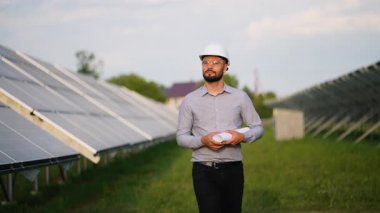 Güneş paneli çiftliğinde yürürken elinde planlar olan kendinden emin erkek mühendis sürdürülebilir yeşil elektrik üretimi için alternatif enerji santralini inceliyor.