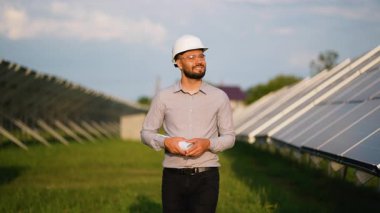 Beyaz şapkalı gülümseyen erkek mühendis bir güneş enerjisi santralinde yürüyor, elinde planlar ve arka planda fotovoltaik paneller olan kameraya bakıyor.