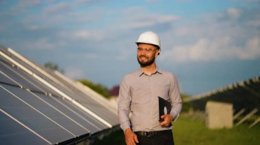 Güneş enerjisi santralinde yürürken elinde dizüstü bilgisayar tutan kendinden emin erkek enerji mühendisi fotovoltaik hücreleri inceleyip kameraya gülümsüyor.