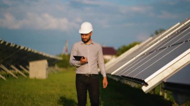Beyaz miğferli bir erkek mühendis elinde tablet, güneş enerjisi santralinde yürüyor, teftiş sırasında fotovoltaik panelleri kontrol ediyor ve gün batımında kameraya gülümsüyor.