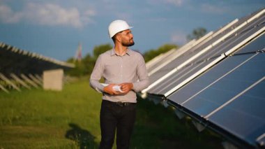 Güneş enerjisi santralinde yürüyen kendinden emin erkek mühendis elinde planlar ve sürdürülebilir enerji üretimi için fotovoltaik panellerin sıralarını inceliyor.
