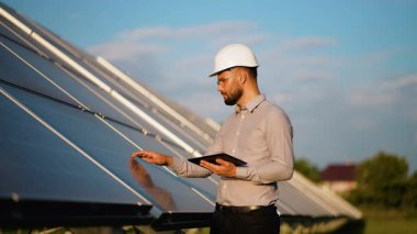 Beyaz şapka ve güvenlik gözlüğü takmış genç erkek mühendis, yenilenebilir enerjinin toplanması ve izlenmesi için bir tablet kullanarak bir enerji santralindeki güneş panellerini kontrol ediyor.