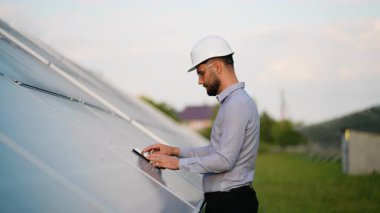 Güneş panelleri arasında duran beyaz başlıklı erkek mühendis tablet üzerindeki sistem verimliliğini kontrol ederek daha yeşil bir gelecek için sürdürülebilir ve yenilenebilir enerji kaynaklarını teşvik ediyor.