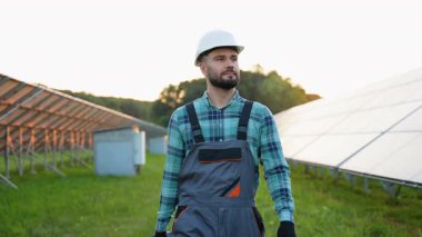 Sert şapkalı ve üniformalı kendinden emin erkek mühendis gün batımında yeşil bir alanda güneş paneli kurulumunu inceliyor, kameraya gülümsüyor. Temiz yenilenebilir enerji ve ekolojik güç kavramı