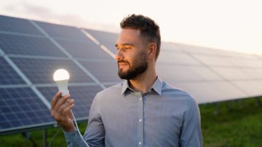 Kendine güvenen sakallı bir adam elinde parlayan bir ampul tutuyor, güneş paneli kurulumunun önünde duruyor, gülümsüyor ve kameraya bakıyor, yeşil enerji kavramını simgeliyor.