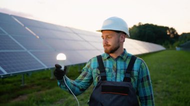 Bir güneş çiftliğindeki fotovoltaik hücrelerden elde tutulan sürdürülebilir elektrik üretimini gösteren bir ampulü tutan sert şapkalı erkek teknisyen, temiz, yenilenebilir enerji kavramını gözler önüne seriyor.