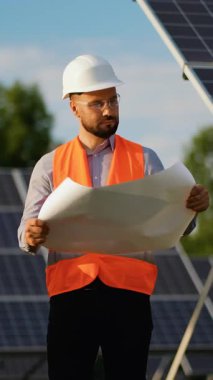 Güneş enerjisi santralinin önünde duran kasketli ve güvenlik yeleği giyen kendinden emin erkek mühendis, bir gülümsemeyle projenin planını açıp inceliyor, sürdürülebilir enerji çalışmaları sergiliyor.
