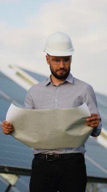 Sağlam şapkalı kendinden emin erkek mühendis elinde bir planla yenilenebilir enerji istasyonundaki güneş panellerini inceliyor. Teknisyen fotovoltaik kurulum projesini kontrol ederken gülümsüyor