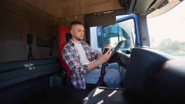 Genç, profesyonel bir erkek sürücü, güneşli bir günde bir kamyon kabininde oturuyor ve akıllı telefonunu kullanıyor, mesajları kontrol ediyor ya da uzun bir yolculuktan sonra sosyal medyada geziniyor.