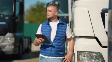 Envanter için dijital tablet kullanan ve bir kargo terminalinde kamyonun yanında durup telsizle konuşan kendinden emin bir erkek sürücü filo ve ulaşım lojistiği yönetiyor.